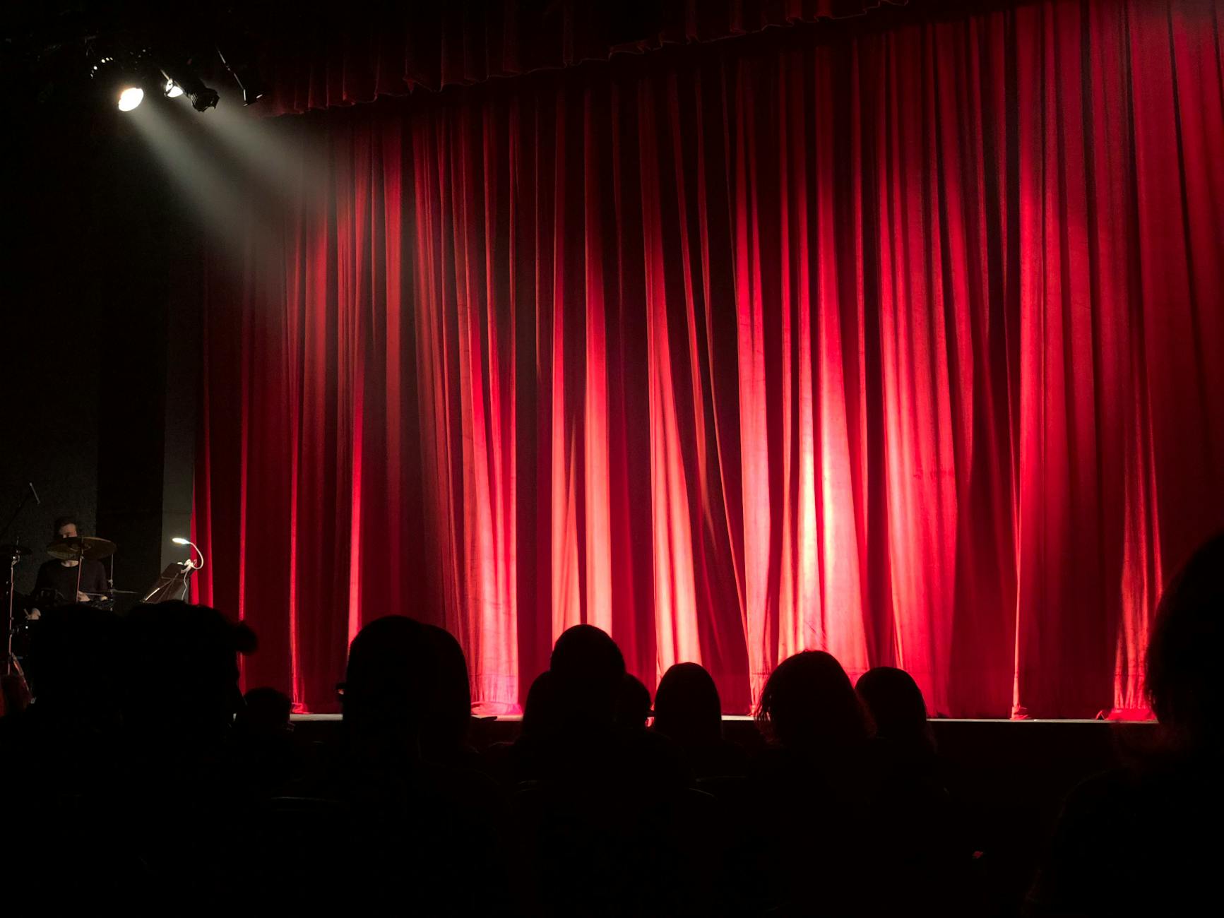 An image of a stage with a red curtain; there are the silhouettes of people's heads in the front rows. Off to the left of the stage is a person sitting behind a drum set.